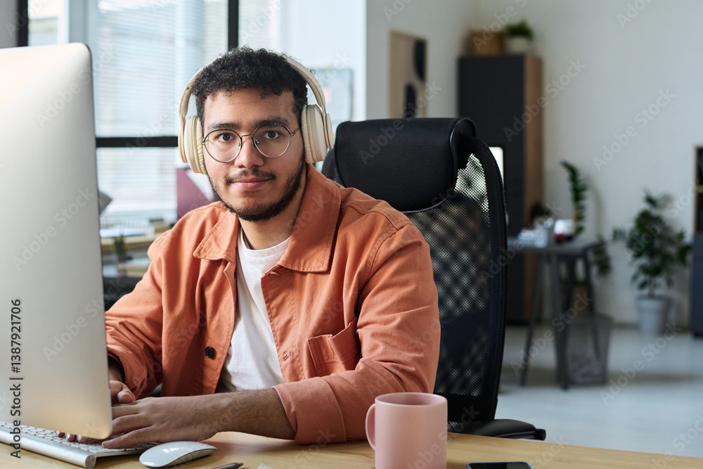 Fototapete Young smiling man in eyeglasses and headphones looking at camera and typing on computer keypad during network
