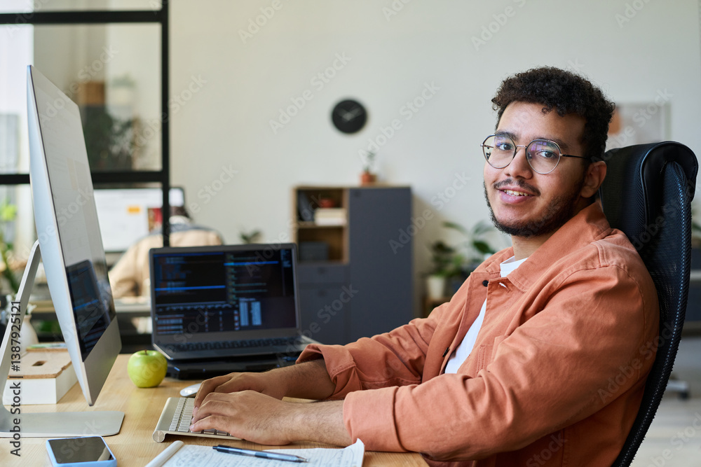 Poster Young smiling male designer or programmer testing new product in front of desktop computer and looking at camera