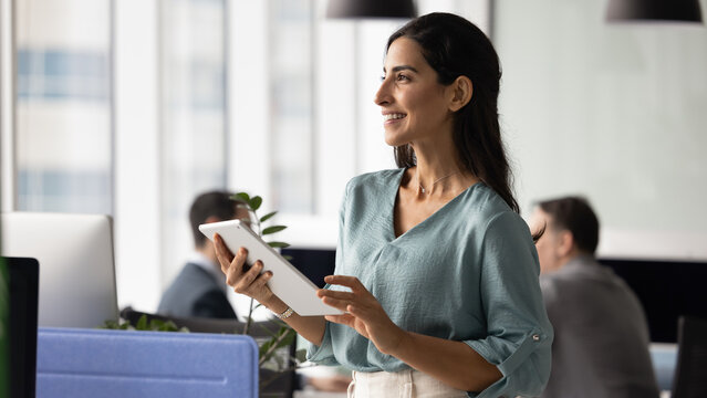 Positive young company leader woman holding digital tablet, standing in office space with colleagues working behind, looking away, smiling, thinking on business plan, Internet marketing. Banner shot