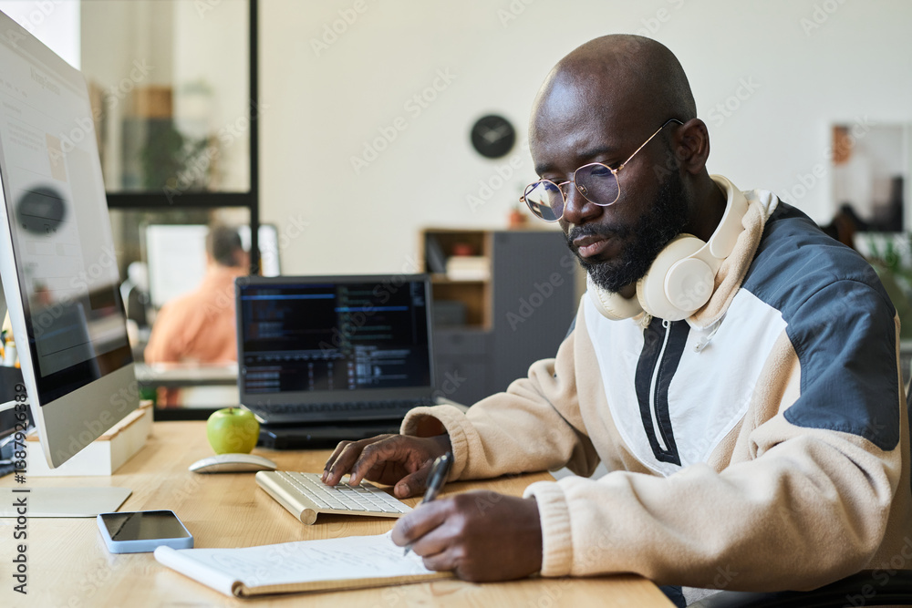 Aufkleber Young serious African American man in casual attire sitting by workplace with desktop computer and taking notes in notebook