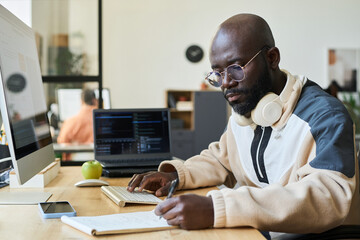 Young serious African American man in casual attire sitting by workplace with desktop computer and taking notes in notebook