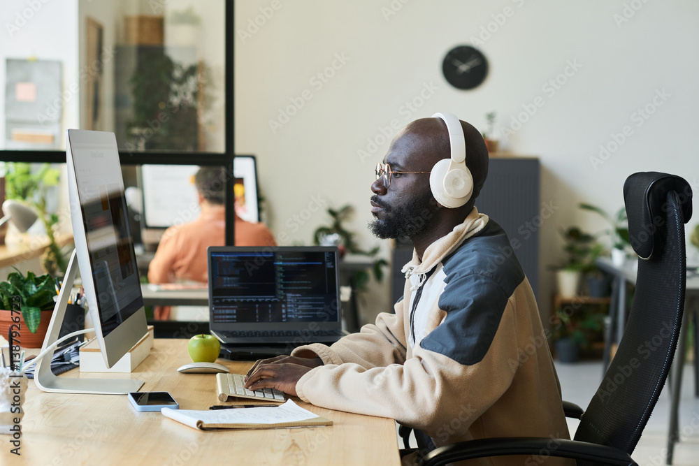 Poster young serious designer in headphones sitting by workplace in front of desktop computer screen with v