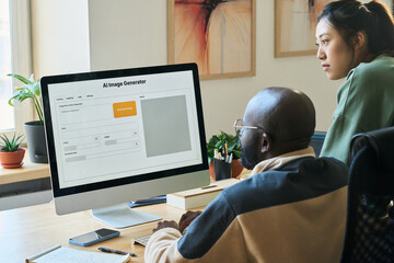 Two young intercultural office workers looking at screen of desktop computer with AI image generator by workplace
