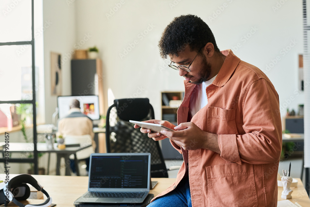Poster Young serious software developer with tablet working over new project while sitting on desk in coworking space