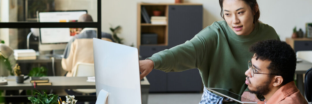 Header of young confident Chinese woman pointing at computer screen while explaining information to multiethnic male colleague