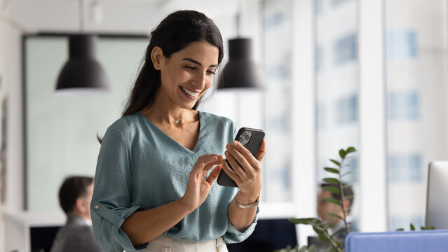 Happy beautiful Hispanic businesswoman using web service on smartphone in office workspace, working on online project on mobile phone, typing on cellphone, smiling. Banner shot - Powered by Adobe