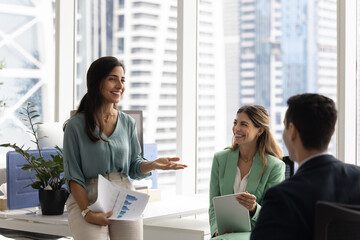 Happy Hispanic business colleagues brainstorming on work project, discussing creative ideas, analyzing financial sales reports, sitting in circle, smiling, laughing. Female leader holding meeting