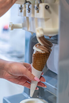 Delicious Treat Preparation: Close-up of female hand holding crispy waffle cone receiving a perfectly formed creamy swirl of mixed chocolate and vanilla soft serve ice cream from dispenser machine.