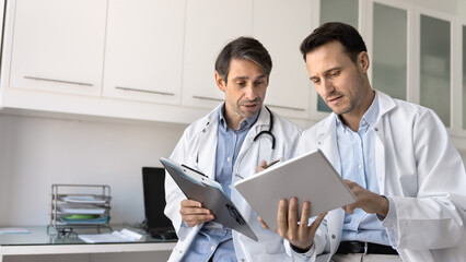 Two serious cardiologists engaged in discussion in modern consulting room, analyze medical reports, evaluating diagnosis or lab results on digital tablet device, share knowledge and opinions. Teamwork
