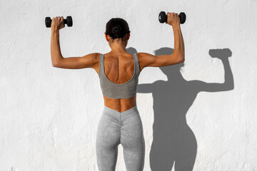 A fit woman in gray athletic wear lifting dumbbells above her shoulders, standing with her back to...