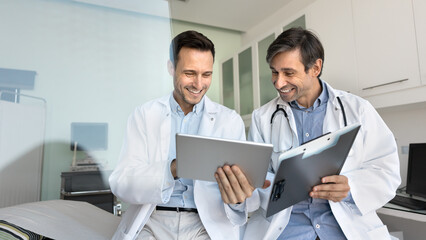 Two smiling male doctors in coats using digital tablet in clinic office, communicating with...