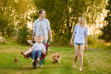 Man walks barefoot on grass next to woman and carries his son and daughter in garden wheelbarrow, dogs run nearby. Gardening concept, country family vacation.