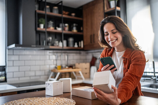 Woman opening package and using smartphone in kitchen