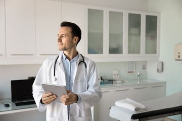 Contemplative Latin doctor dressed in white coat holding tablet, gazes thoughtfully into distance, standing in modern medical consulting room. Digital telemedicine tools usage, innovations, workflow