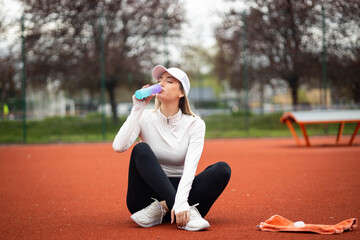 A young woman taking a refreshing sip from her water bottle while seated on a tennis court, showcasing her active lifestyle.