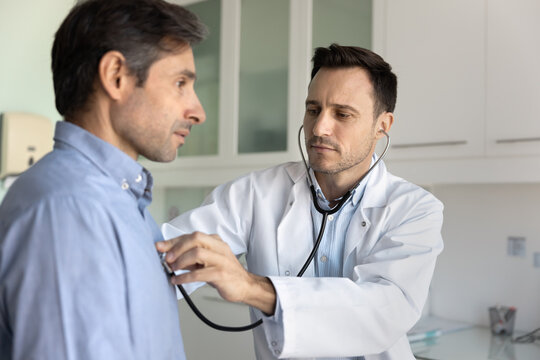 Middle-aged 40s cardiologist using stethoscope to listen to male patient heartbeat, assessing cardiac or respiratory health during medical examination in consulting room. Cardiology, professional care