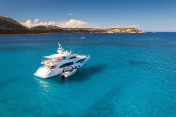 Aerial view of luxury yacht on blue sea at susnet in summer. Sardinia, Italy. Tropical seascape with speed boat, yacht, sea lagoon, mountain, transparent azure water, sky with clouds. Top drone view
