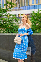 Fototapeta premium A woman in a blue polka dot dress is standing in front of a building. She is holding a white purse and smiling