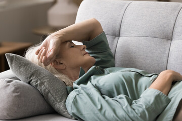 Pause to relax. Portrait elderly woman retiree lying down on sofa with closed eyes hold hand on...