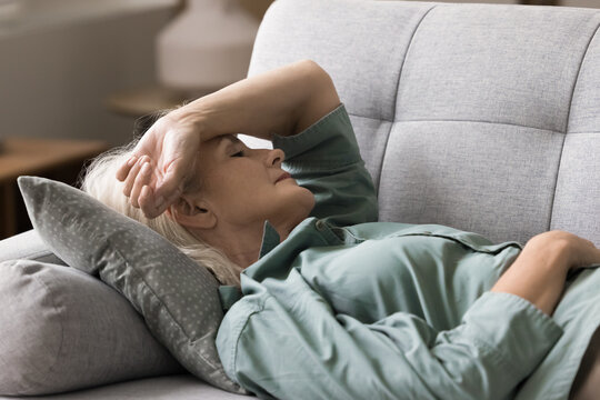 Pause to relax. Portrait elderly woman retiree lying down on sofa with closed eyes hold hand on head take sleep nap to rest feel weak dizzy tired fatigue find moment of relief from menopause symptoms - Powered by Adobe