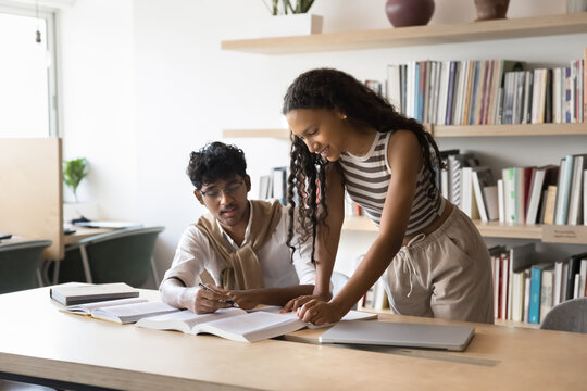 Friendly peer learning. Two young diverse multiethnic students mates engaged in self studying at library discuss books review scientific literature. Teen girl help male groupmate in writing coursework