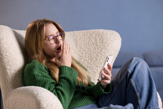 Yawning woman in glasses looks at phone, showing signs of digital fatigue and sleepiness.