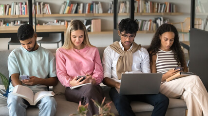 Modern study session. Diverse group of multiethnic teen students sit in row on sofa at campus library use mix of gadgets paper materials for learning self training involved in personal education tasks