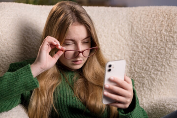 Girl in green sweater struggles to read smartphone, adjusting glasses with puzzled look.