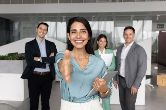 Happy Latin business leader woman offering handshake at camera, posing for portrait with group of colleagues in background, welcoming for hiring, career start, joining successful team