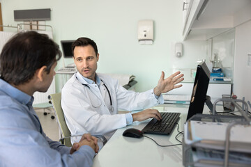 Obraz premium Male doctor sits in workplace with computer explaining information, engaged in conversation about health concerns, potential treatments, or preventive care measures to patient. Medical consultation
