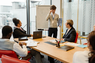 A group of five employees are sitting at a table and smiling while looking at a male intern who is standing next to them and laughing while holding a clipboard, in an office, during an internship