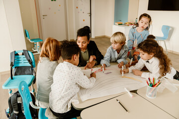 A group of five schoolchildren and a teacher sit and stand at a table while drawing, at school, in a classroom