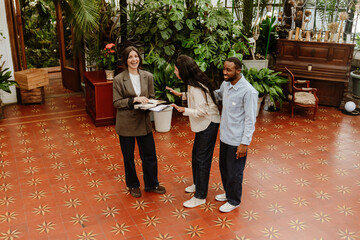 A man stands and holds his hand behind the back of a woman in front of him and looks at a woman organizer who is holding a clipboard and a notepad, they are laughing while preparing for the wedding