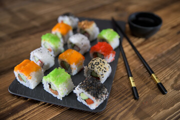 Colorful assorted sushi rolls arranged on a dark slate plate with chopsticks and soy sauce on wooden table