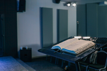 Open Bible resting on a black podium inside a modern church hall, with chairs and musical instruments visible in the background