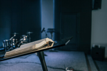 Open Bible on a black podium in a dimly lit church auditorium with blurred musical instruments in...