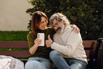 A man is sitting on a bench and showing a thumbs up while looking at the phone held by a woman...