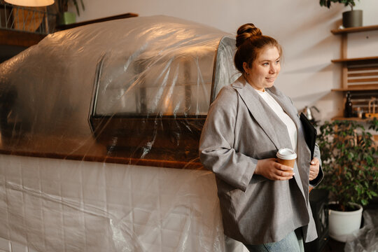 Female owner standing holding a cup and computer tablet while leaning on the bar and smiling, preparing to open the cafe