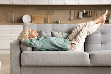 Giving rest for legs. Serene smiling older woman spend leisure time lying on back on comfy sofa at...