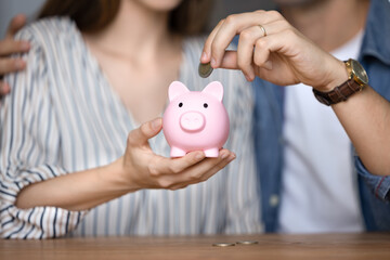 Close up cropped view of unknown married couple drop coins into small pink piggy bank. Young family...