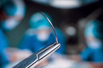 Hemostatic forceps holding half circle cutting needle and suture in front of of blurry operators under the lights in operating room. Surgical team working in an operation room.