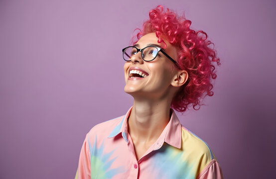 Happy non-binary person with vibrant pink curly hair wears glasses in studio portrait. Smiling person with bright, colourful tie dye shirt celebrates freedom, diversity, individuality. Positive vibes.