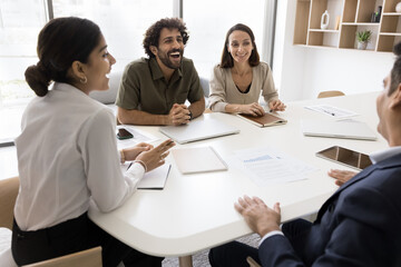 Four multinational workmates attend group briefing gathered together around table, laughing, joking, having good friendly work relationships, share creative ideas and solutions in modern meeting room