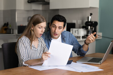 Married couple sit at table in kitchen, analyzing financial documents with surprised or concerned...