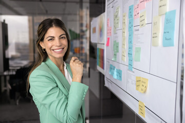 Cheerful young Hispanic company CEO woman using scrum management method at Kanban board, looking at...