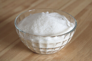 Glass bowl overflowing with white granulated sugar on wooden table
