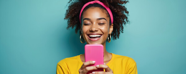 Happy young woman holding pink smartphone, laughing at screen. African female with curly hair, casual yellow t-shirt. Modern mobile communication technology. Joyful face expression.