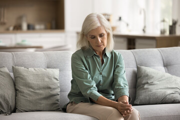 Lack of purpose. Calm uncertain mature woman spend time at home sitting alone on sofa lost in...