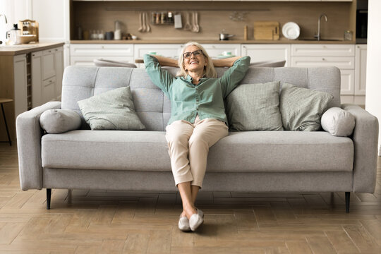 Finding peace at cozy home. Serene tranquil older woman homeowner renter tenant enjoy moment of relax on comfy sofa listen to music feel peace of mind in clean light interior of large studio apartment