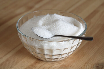 Glass bowl overflowing with white sugar and spoon on wooden table